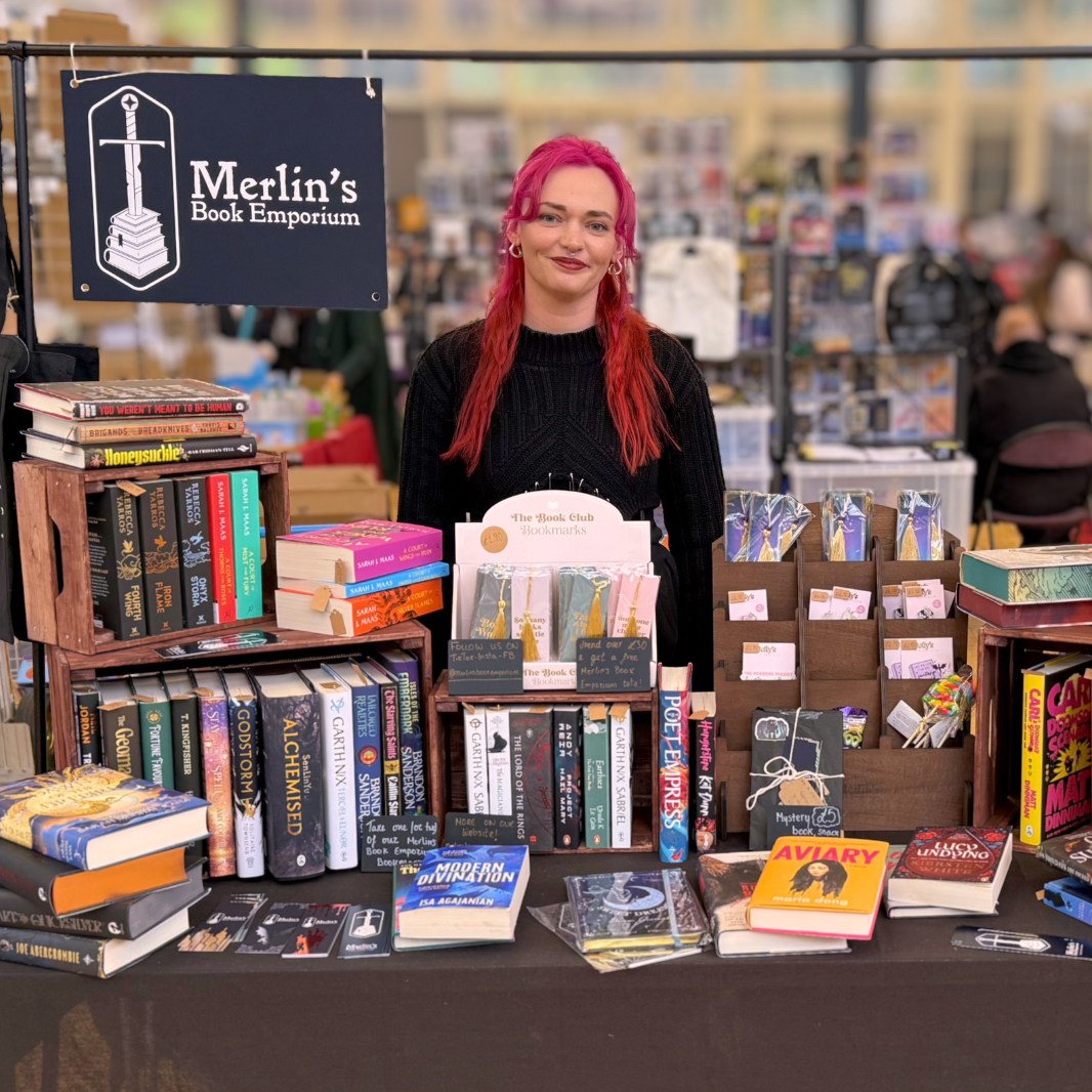 Person with red hair standing behind a table filled with books at 'Merlin's Book Emporium'.