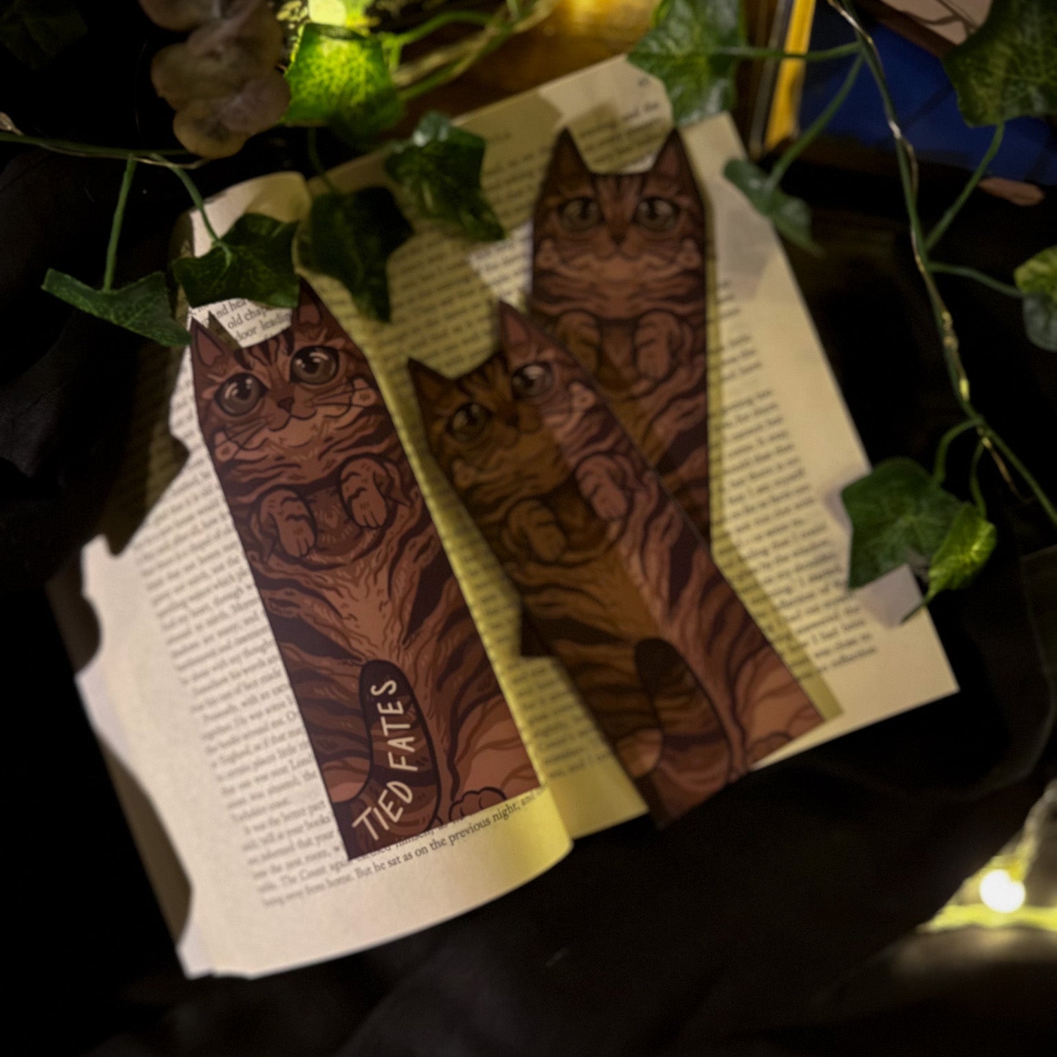 Bookmarks with cat illustrations on an open book, surrounded by greenery and string lights.