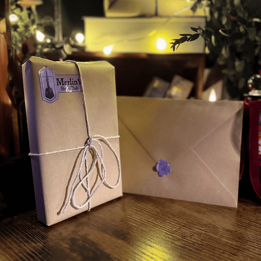 A wrapped package tied with twine and a matching envelope with a purple wax seal, styled on a wooden surface with warm string lights in the background. A label on the package reads 'Merlin's'.