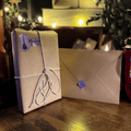 A wrapped package tied with twine and a matching envelope with a purple wax seal, styled on a wooden surface with warm string lights in the background. A label on the package reads 'Merlin's'.